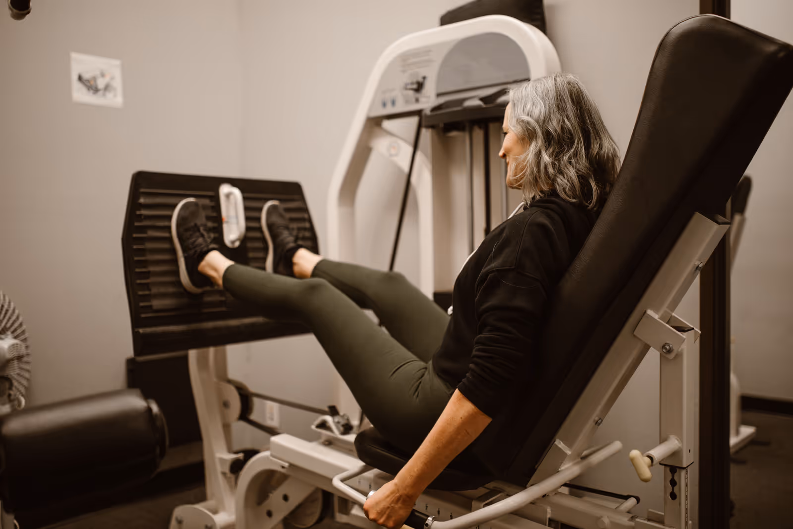 Mulher de cabelos grisalhos usando um aparelho de leg press em uma academia, pernas apoiadas na plataforma de peso